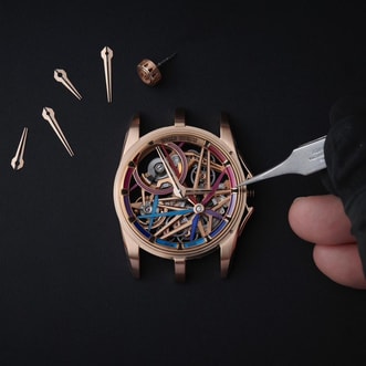 This image shows a hand holding fine tweezers putting the finishing touches on the components of a Roger Dubuis MONOBALANCIER Blacklight Spin-Stone™ watch. The watch's hands and crown are positioned around the case.