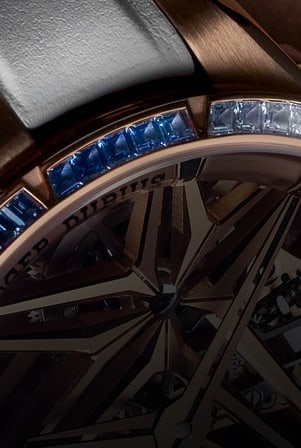 The image is a detailed close-up of the Roger Dubuis Excalibur Monotourbillon RDDBEX1134 watch, highlighting the rail setting of its bezel. It shows blue sapphires and baguette-cut diamonds aligned in a rose gold case, with part of the skeletonized dial and the Maison name visible.