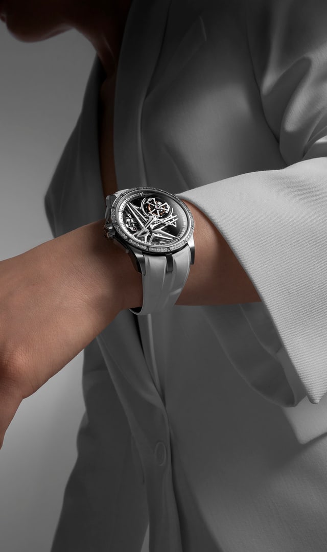 The image shows the Roger Dubuis Excalibur Monotourbillon RDDBEX0989 watch worn on the wrist of a person wearing a white jacket, in an elegant and minimalist setting.
