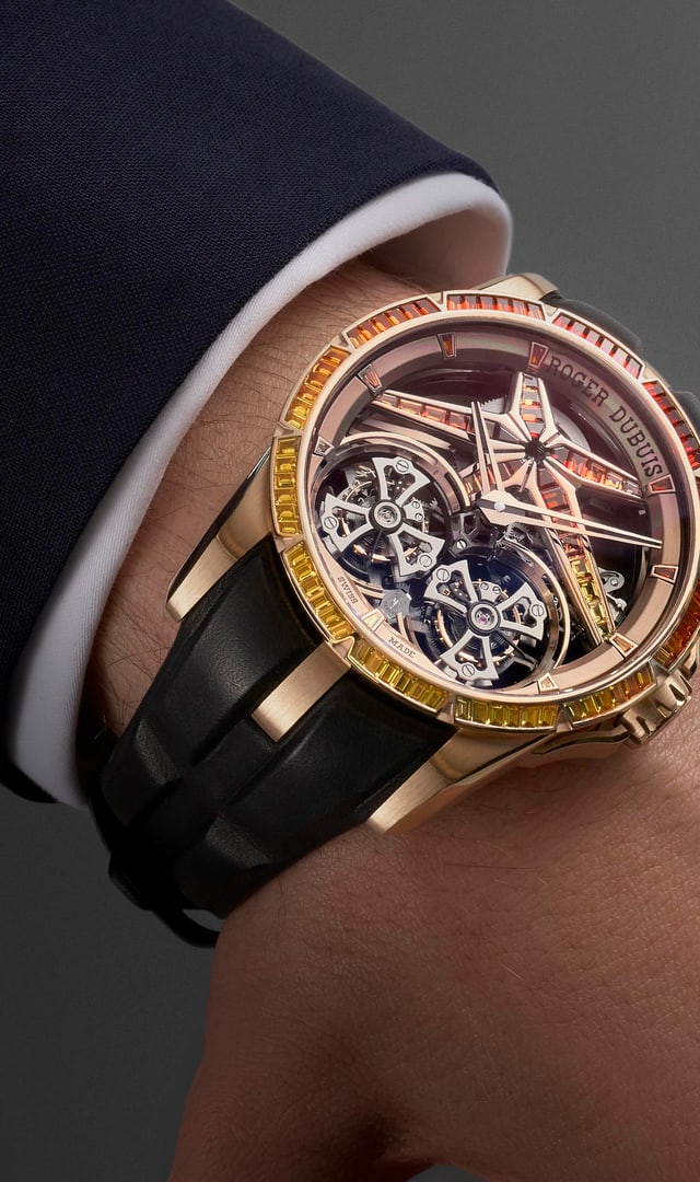 The image shows the Roger Dubuis Excalibur Double Tourbillon RDDBEX1130 watch on the wrist of a person wearing a dark suit and white shirt. The watch features a gold case, a black strap, and a skeletonized dial adorned with warm-colored gemstones (yellow, orange, red), highlighting its intricate mechanism.