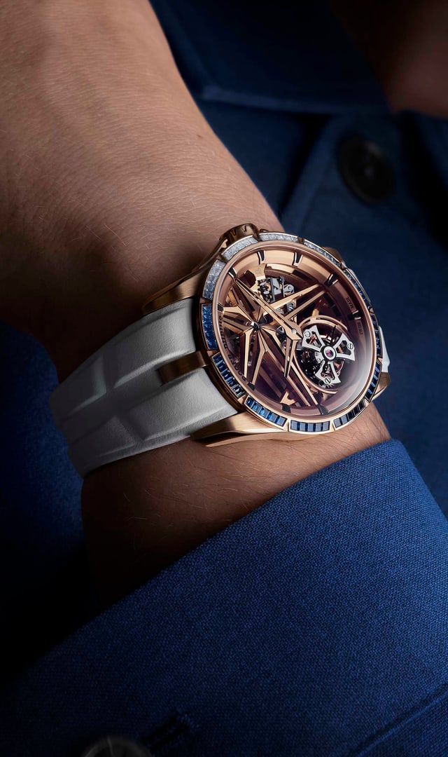 The image shows the Roger Dubuis Excalibur Monotourbillon RDDBEX1134 watch on the wrist of a person wearing a blue shirt. The rose gold case, the bezel set with blue sapphires and diamonds, and the white strap are visible in an elegant, slightly dark setting.