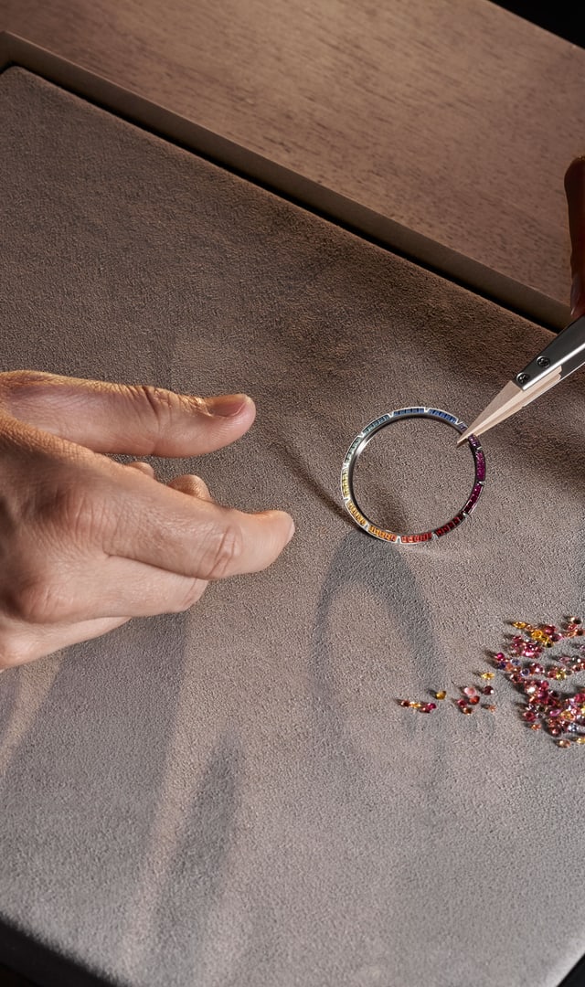 The image features two bezels set with precious stones, a bezel set with stones, individual stones (emeralds, diamonds, sapphires) and part of a diamond-encrusted bracelet, all arranged on a grey surface with tweezers.