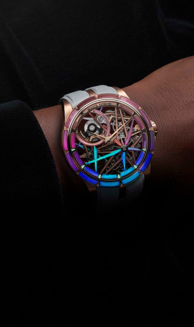 The image shows a close-up of a wrist wearing the Roger Dubuis Excalibur Monobalancier Blacklight RDDBEX0950 watch, characterized by its skeletonized dial adorned with phosphorescent synthetic stones in neon shades of pink, purple, and blue, which stand out against a dark background.